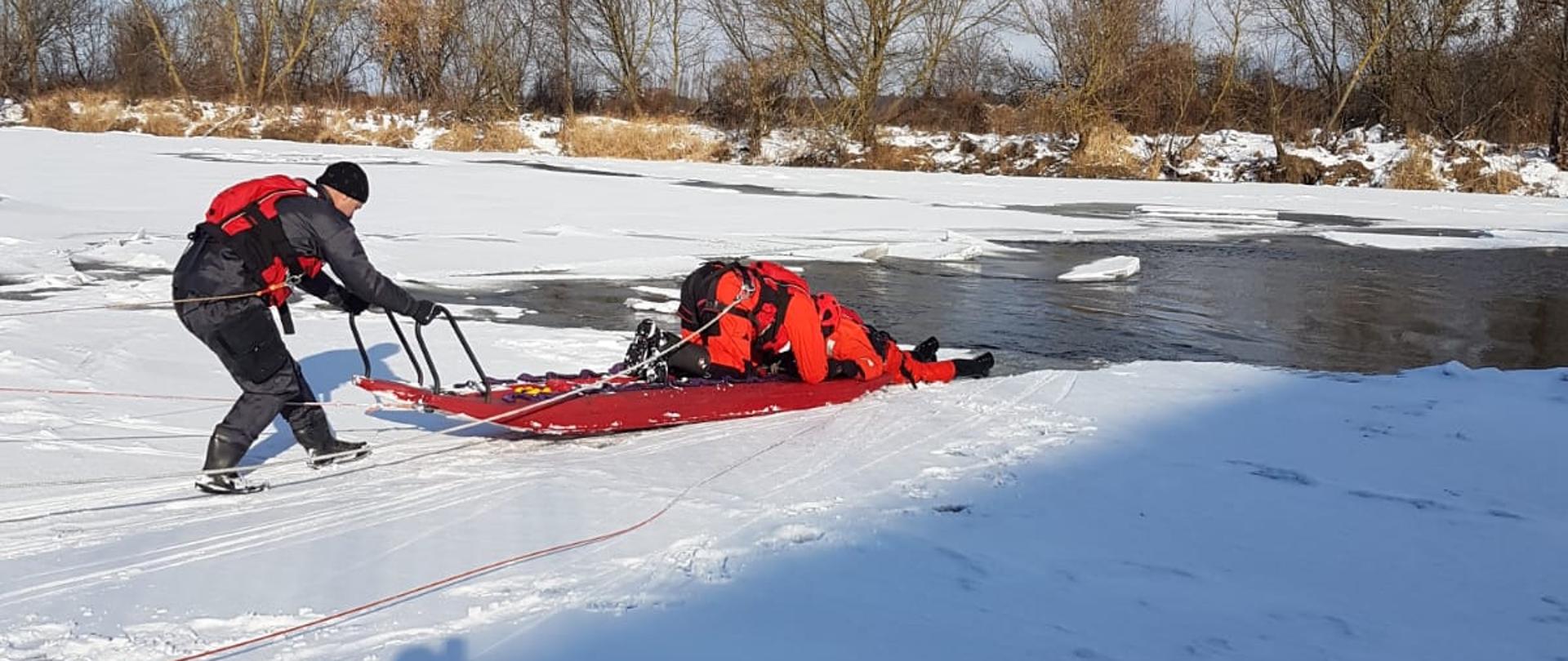 Dwóch ratowników na zamarzniętym akwenie przy pomocy sań lodowych podejmuje próbę wydobycia poszkodowanego na brzeg.