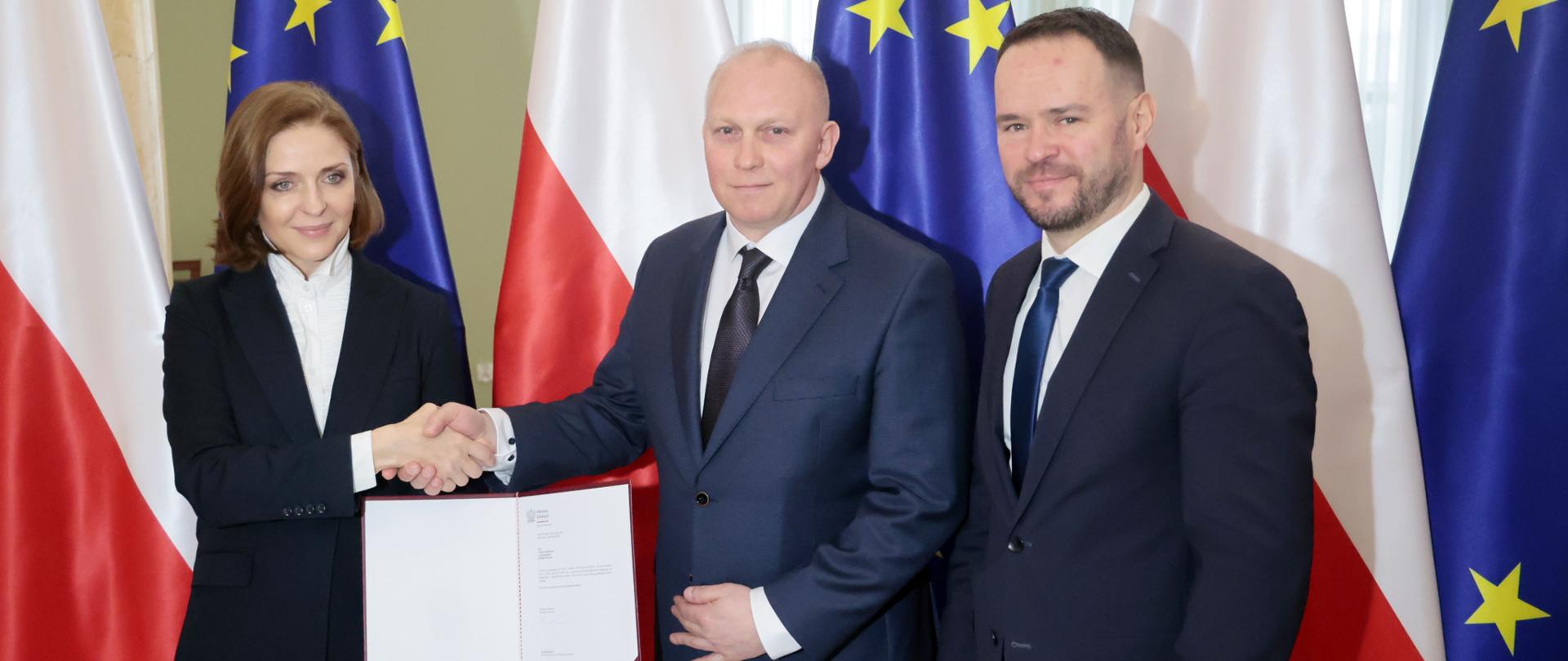 Kobieta podaje rękę mężczyźnie w garniturze jednocześnie trzymając w drugim ręku otwarty dokument. Obok nich stoi mężczyzna w garniturze, w tle flagi Polski i Unii Europejskiej. 