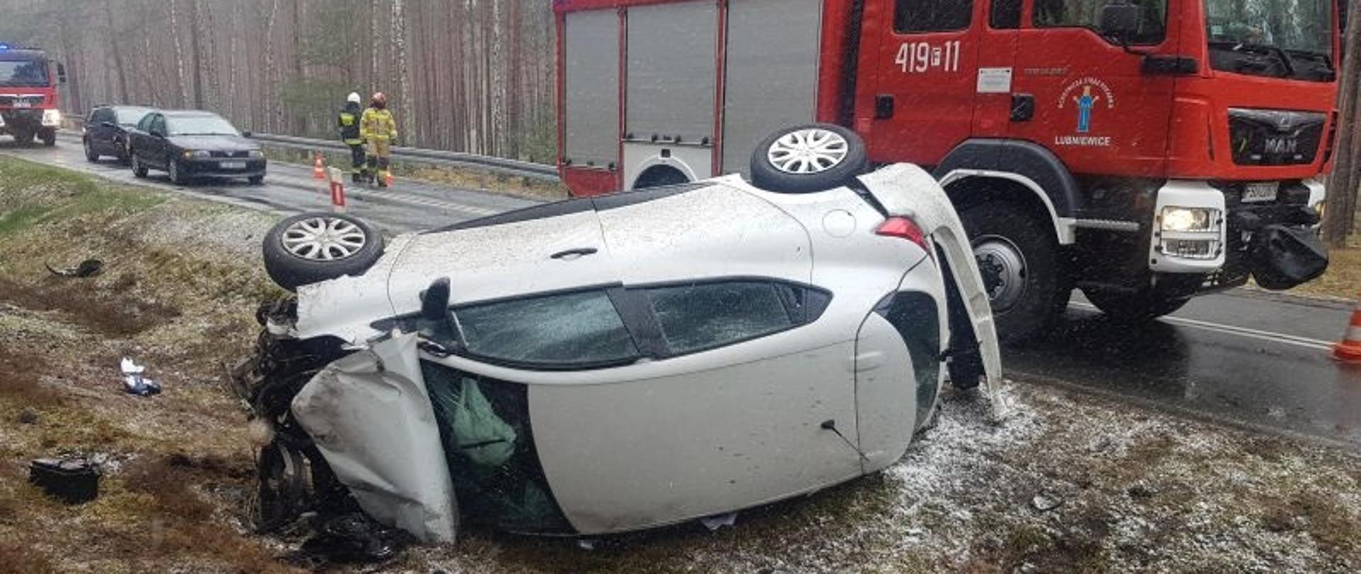 zdjęcie przedstawia pojazd osobowy Renault Clio leżący na boku na poboczu, w tle widać pojazdy straży pożarnej oraz ratowników 
