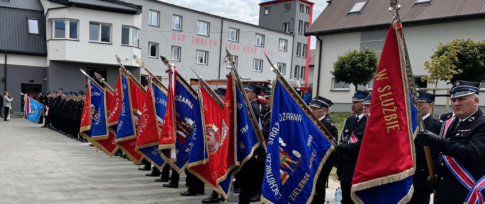 plac przed budynkiem komendy z napisem. Po prawej stronie kadru, wzdłuż placu, stoi długi szpaler pocztów sztandarowych jednostek Ochotniczych Straży Pożarnych, trzymających rozpostarte, ozdobne czerwone i niebieskie sztandary ze złotymi frędzlami. Na jednym z niebieskich sztandarów widnieje napis „OCHOTNICZA STRAŻ POŻARNA W ZIELONCE NOWEJ”. W głębi wybrukowanego placu uformowany jest pododdział strażaków, a przy bramach garażowych zaparkowane są wozy strażackie oraz czerwony samochód osobowy.