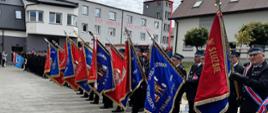 plac przed budynkiem komendy z napisem. Po prawej stronie kadru, wzdłuż placu, stoi długi szpaler pocztów sztandarowych jednostek Ochotniczych Straży Pożarnych, trzymających rozpostarte, ozdobne czerwone i niebieskie sztandary ze złotymi frędzlami. Na jednym z niebieskich sztandarów widnieje napis „OCHOTNICZA STRAŻ POŻARNA W ZIELONCE NOWEJ”. W głębi wybrukowanego placu uformowany jest pododdział strażaków, a przy bramach garażowych zaparkowane są wozy strażackie oraz czerwony samochód osobowy.
