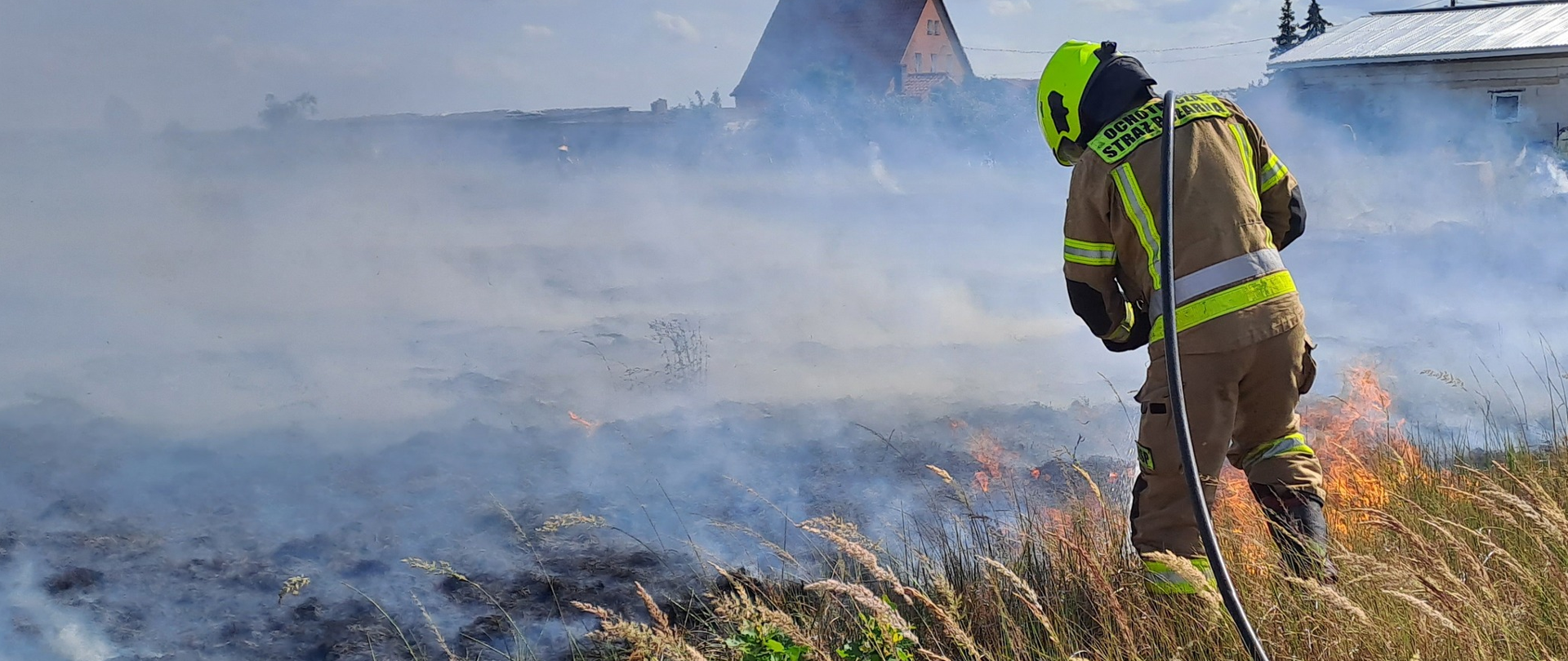 Strażak gaszący pożar traw. W tle widoczna jest spalona łąka, nad którą unosi się dym. Zdjęcie powiększy się po kliknięciu na nie.
