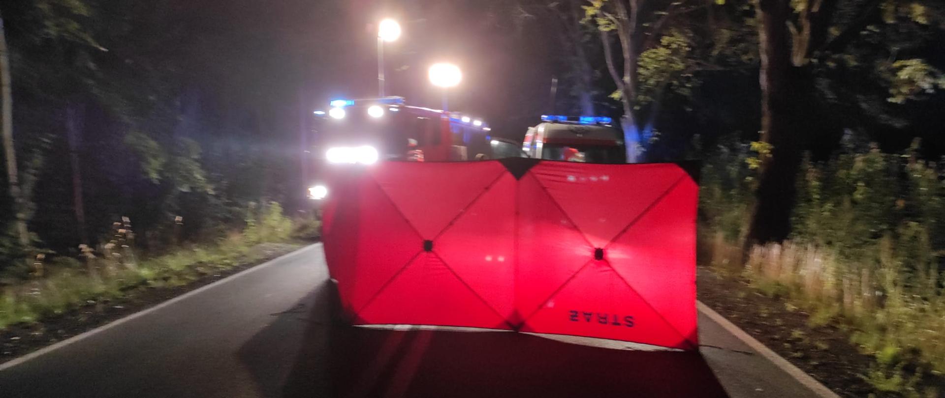 Zdjęcie zrobione nocą. Czerwony parawan na drodze. Światła samochodu strażackiego oświetlają miejsce zdarzenia.