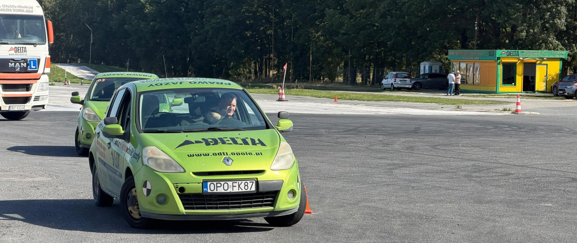 Jasnozielony samochód osobowy marki Renault z logo ośrodka jazdy wykonuje skręt wokół pachołka na asfaltowym placu manewrowym. Za kierownicą widoczny jest kierowca. W tle po lewej stronie stoi ciągnik siodłowy, a po prawej żółty budynek ODTJ. Ćwiczenie w ramach kategorii B.