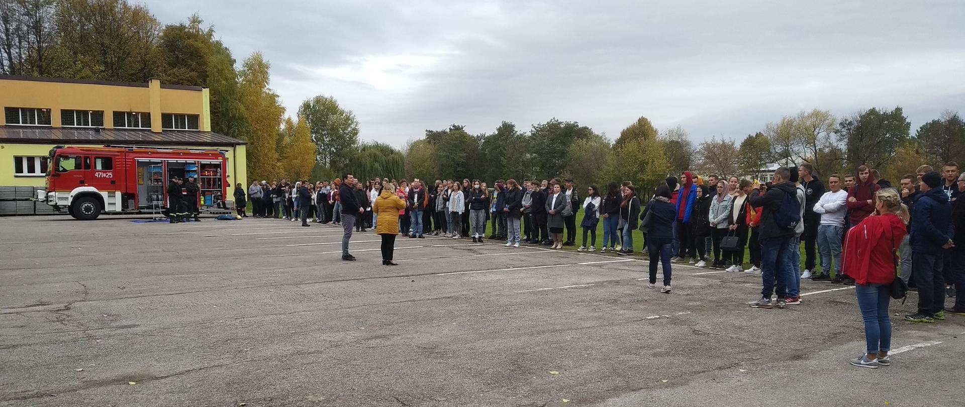 Zdjęcie wykonane na placu szkolnym. Przedstawia zbiórkę młodzieży szkolnej po przeprowadzonej ewakuacji. Po lewej stoi samochód strażacki z otwartymi skrytkami i widać sprzęt strażacki. Trzech strażaków obserwuje młodzież.