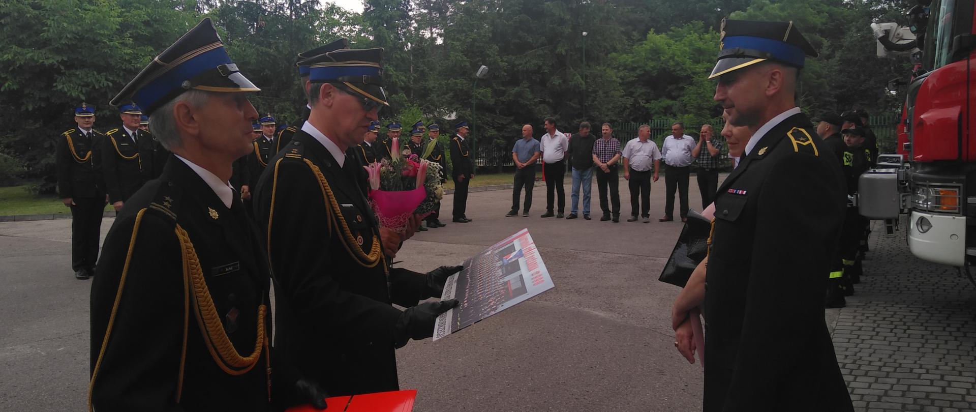 Strażacy stoją naprzeciw siebie. Jeden ze strażaków trzyma w rękach zbiorową fotografię z zamiarem jej wręczenia strażakowi stojącemu naprzeciwko. Drugi ze strażaków trzyma czerwony skoroszyt, który jest opasamy złotą wstążką. W oddali strażacy oraz emeryci pożarnictwa ustawieni w szeregu stoją na zbiórce. 