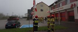 Strażacy podczas zmiany służbowej przed budynkiem JRG w Miechowie w umundurowaniu bojowym dokonują uroczystego wciągnięcia flagi na maszt. 