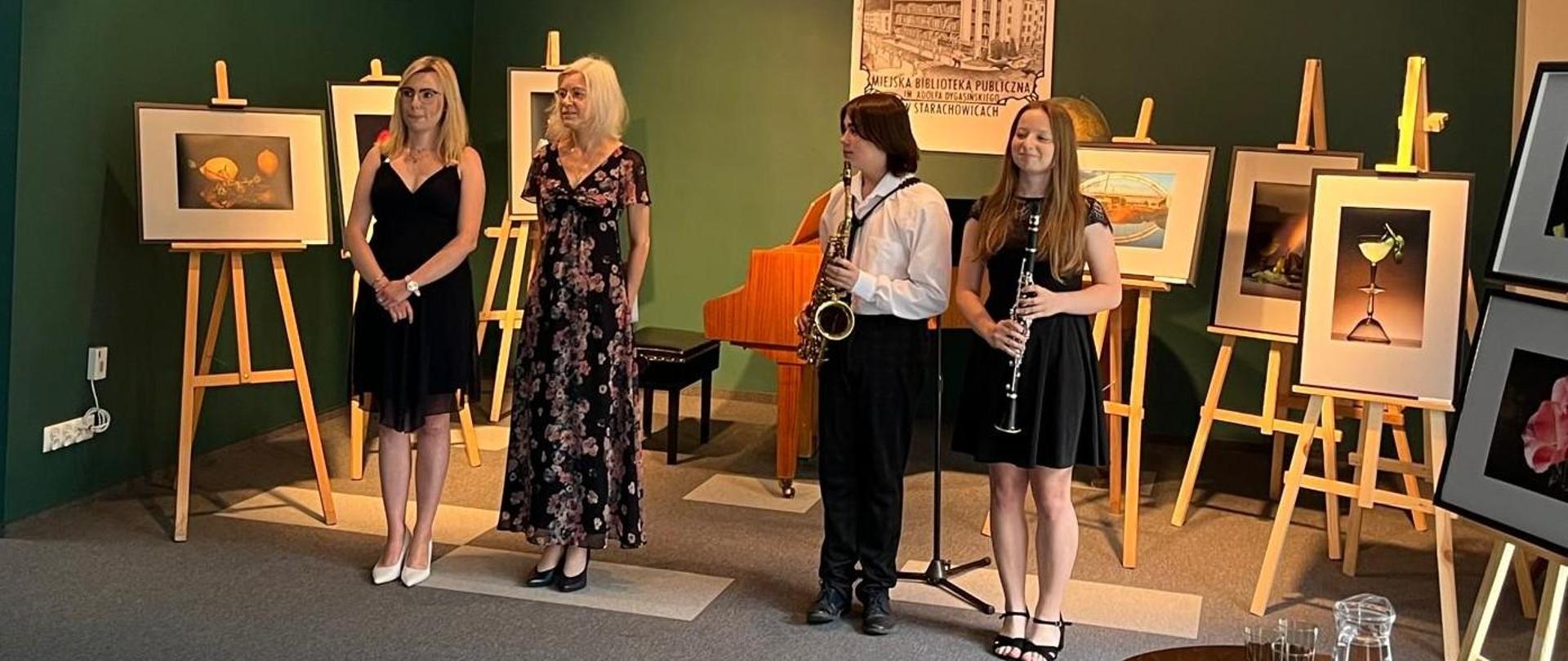 Na zdjęciu i widać uczestników koncertu którzy stoją przed sztalugami na których widnieją obrazy w tle widoczny fortepian oraz ciemnozielona ściana