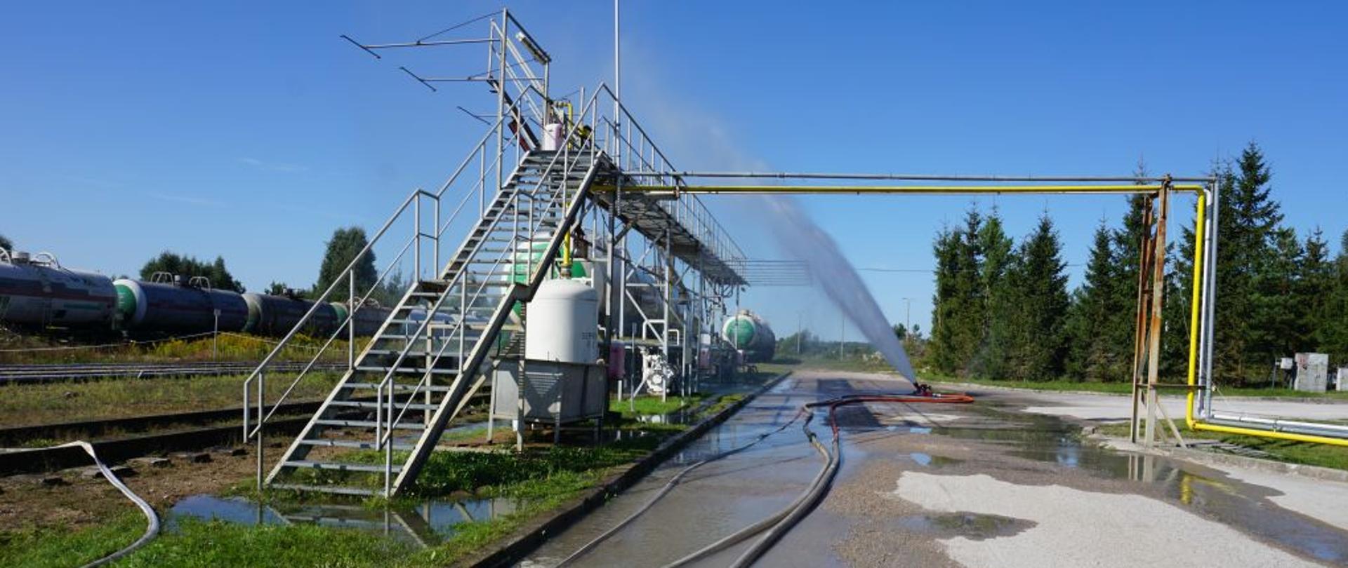 Działko wodne podaje prąd wody na cysternę kolejową. Do działka prowadzą dwa węże strażackie.