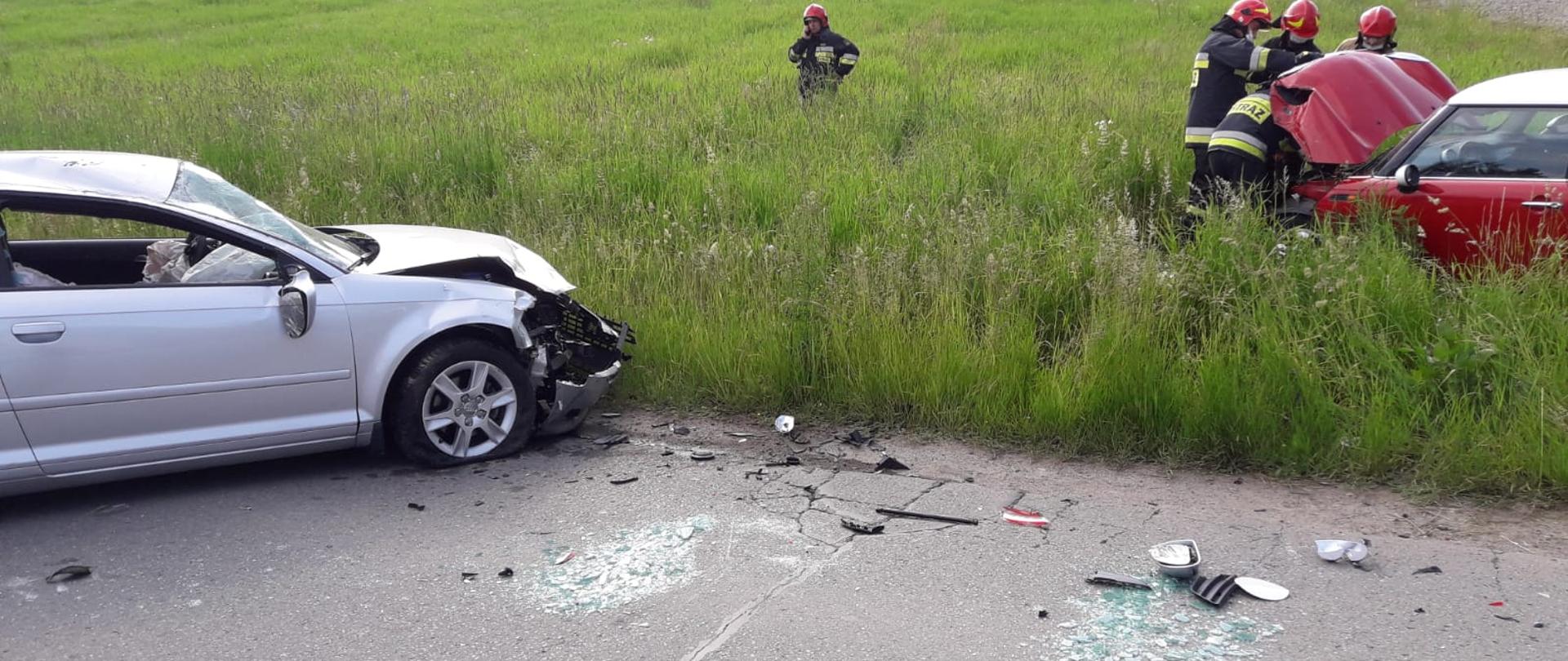 Połowa jasnego samochodu osobowego z uszkodzonym przodem oraz fragment samochodu osobowego koloru czerwonego z otwartą klapą silnika przy której stoją strażacy.