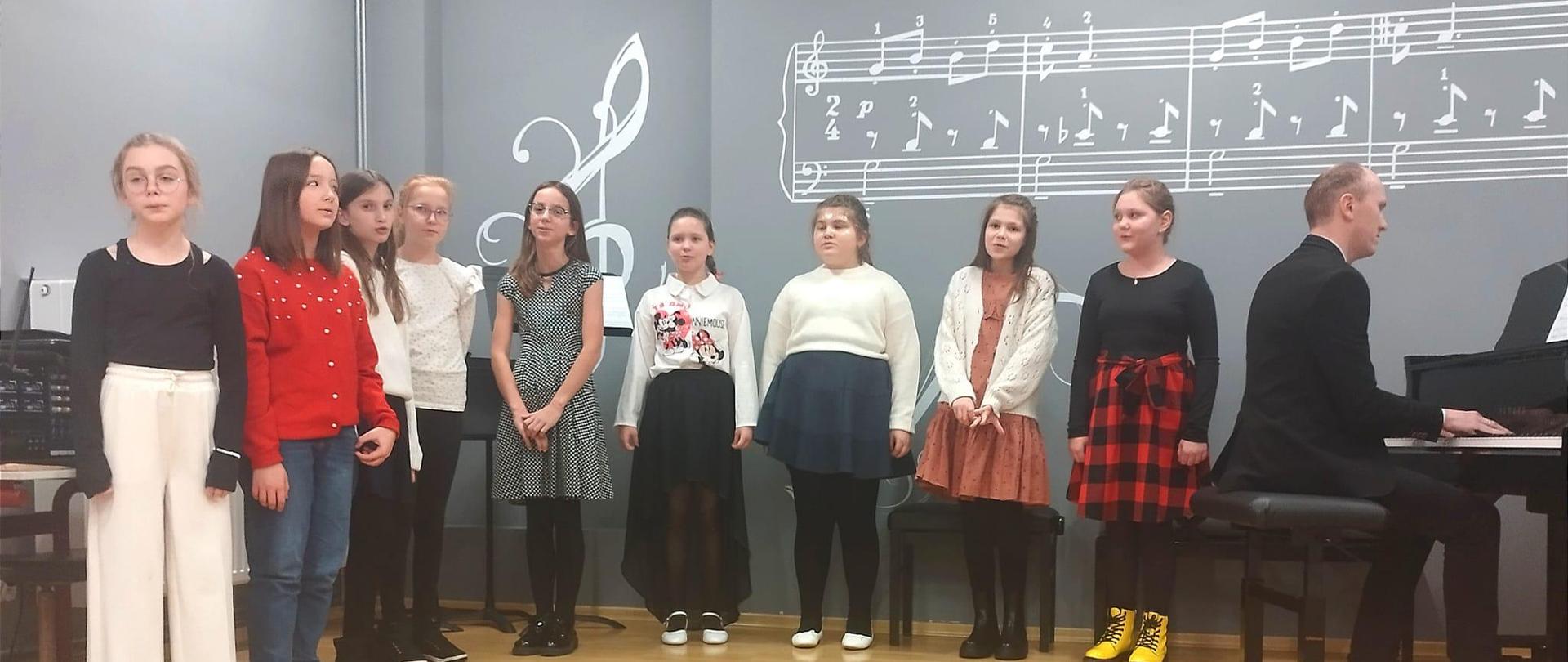 Popis uczniów klasy II i IV. Uczniowie stoja na scenie auli szkoły tło jest koloru szarego po prawej stronie widać fortepian oraz nauczyciela 