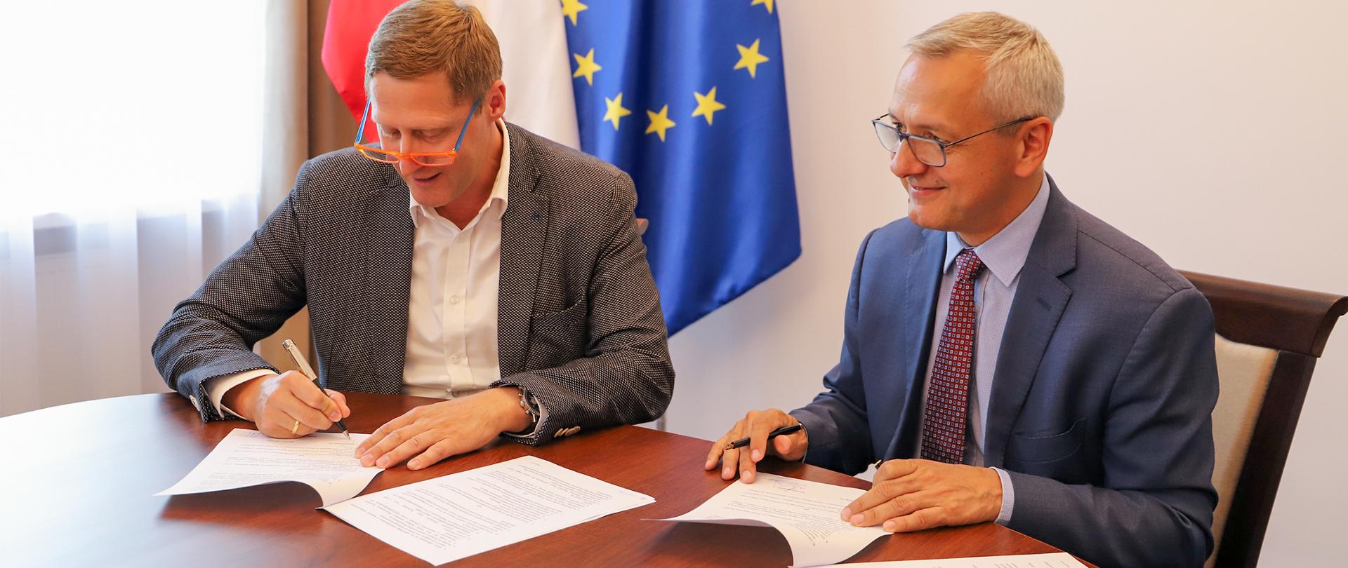 Dwóch mężczyzn siedzących przy stole podpisuje dokumenty. W tle flagi polska i unijna.