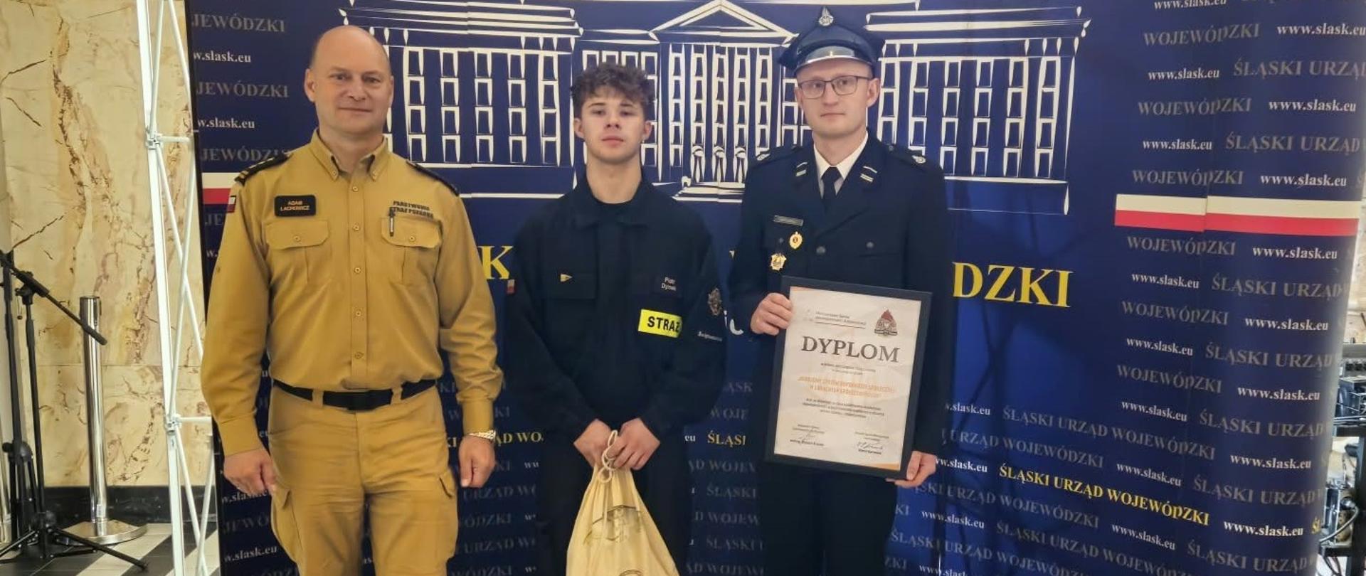 Na zdjęciu przedstawiono uroczystość wręczenia dyplomu/wyróżnienia na tle ciemnoniebieskiej ścianki Śląskiego Urzędu Wojewódzkiego: Osoba po lewej stronie: Mężczyzna w piaskowym/beżowym koszulowym mundurze Państwowej Straży Pożarnej (PSP) z identyfikatorem imiennym (ten sam funkcjonariusz, który na poprzednim zdjęciu stał po prawej stronie). Osoba w środku: Młody mężczyzna w czarnym mundurze strażackim z naszywką „STRAŻ”, trzymający w rękach jasny worek/plecak upominkowy. Osoba po prawej stronie: Mężczyzna w wyjściowym, ciemnogranatowym mundurze strażackim z czapką garnizonową (rogatywką/czapką wyjściową), trzymający oprawiony w ramkę dyplom. Otoczenie i tło:W tle znajduje się ta sama banerowa ścianka Śląskiego Urzędu Wojewódzkiego z grafiką budynku oraz adresami strony internetowej. Całość odbywa się w tym samym wnętrzu z posadzką w czarno-białą szachownicę. 