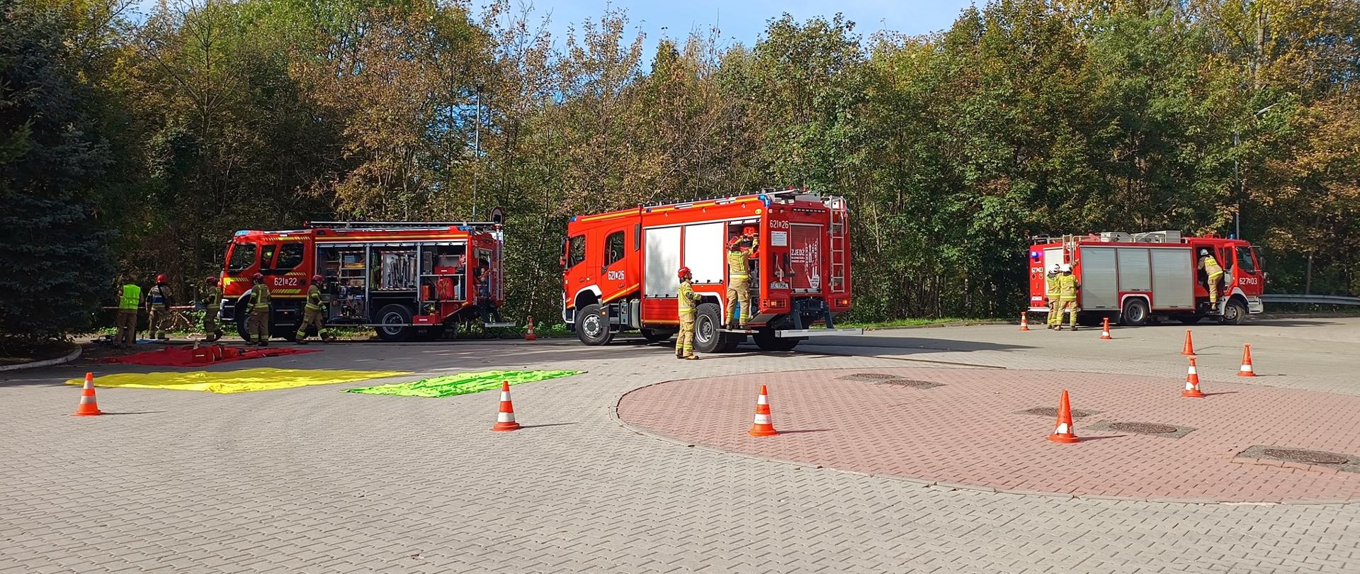 Zdjęcie przedstawia trzy czerwone wozy strażackie zaparkowane w rzędzie na przestronnym placu wyłożonym kostką brukową. Pojazdy mają otwarte rolety schowków sprzętowych, a wokół nich pracują strażacy w piaskowych mundurach bojowych i żółtych kaskach. Przed pojazdem po lewej stronie rozłożone są na ziemi kolorowe maty segregacyjne w odcieniach czerwonym, żółtym i zielonym. Na obszarze manewrowym rozstawiono pomarańczowe pachołki ostrzegawcze, wyznaczające strefę działań. Tło sceny stanowią gęste, zielono-żółte drzewa, a całe zdarzenie odbywa się na zewnątrz w słoneczny dzień pod bezchmurnym niebem. 