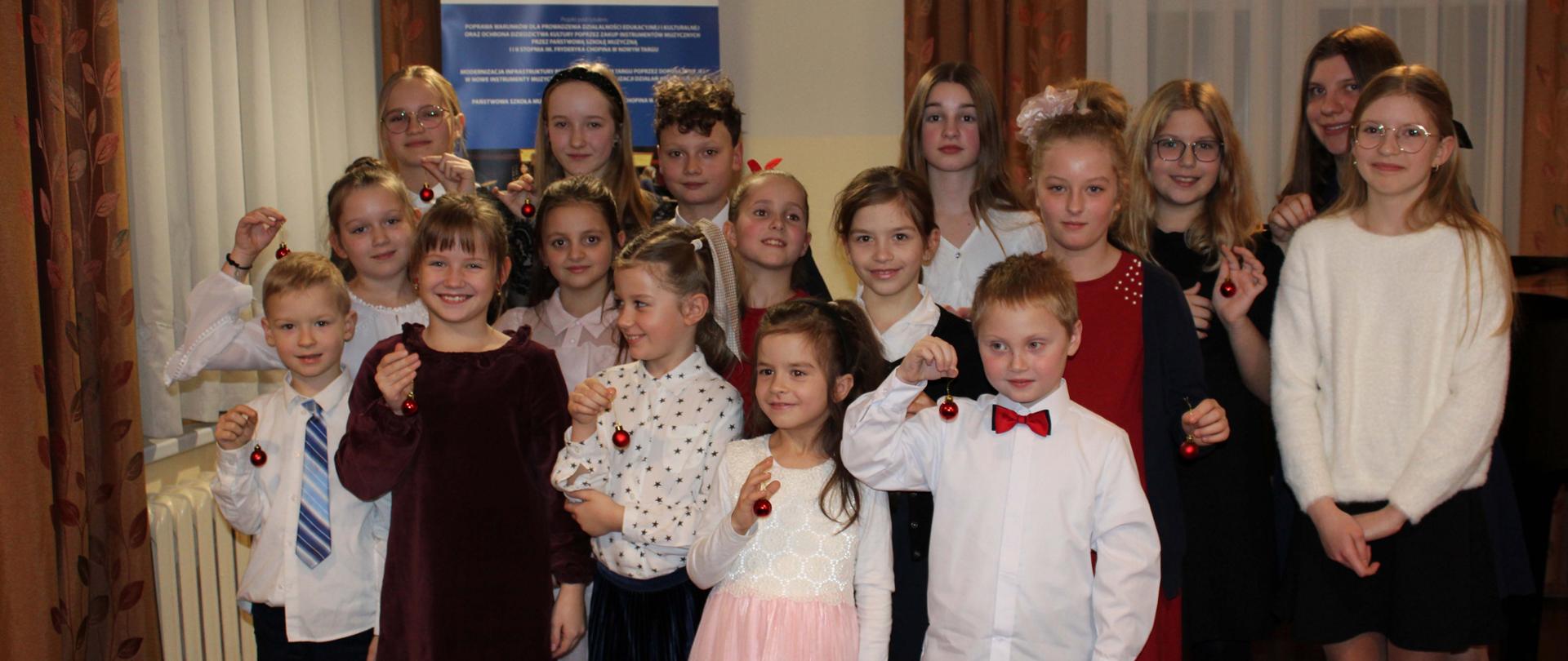 Zdjęcie grupowe. Uczestniczące dzieci w audycji stoją w trzech rzędach. Dzieci uśmiechnięte trzymają w jednej ręce małą czerwoną bąbeczkę podniesioną do góry. Nad nimi dwie zapalone zwisające lampy. Z lewej strony okno z firaną i dwoma zasłonami. Pod oknem kaloryfer. Za dziećmi baner unii europejskiej i drugie okno z firankami i zasłonami