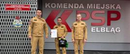 Trzech strażaków w mundurach stoi przed wejściem do komendy miejskiej państwowej straży pożarnej w Elblągu. Strażak stojący pośrodku trzyma w ręku dyplom.