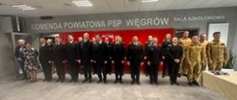 Na zdjęciu widoczna jest grupa funkcjonariuszy Państwowej Straży Pożarnej oraz osób cywilnych zgromadzonych w sali szkoleniowej Komendy Powiatowej PSP w Węgrowie. Uczestnicy stoją w jednym rzędzie na tle czerwonej ściany, pod napisem identyfikującym Komendę. Strażacy występują zarówno w umundurowaniu wyjściowym, jak i służbowym. Po prawej stronie widoczny jest stół z przygotowanymi elementami uroczystości. Całość ma oficjalny i podniosły charakter.