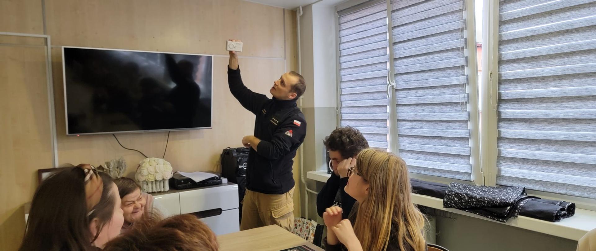 Strażak pokazuje słuchaczom jak działa czujka dymu i czadu.