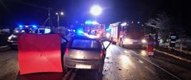 Śmiertelny wypadek w Parszowie. Noc. Na pierwszym planie widoczny tył jednego z samochodów biorących udział w wypadku, parawan. Na dalszym palnie widoczne samochody strażackie, policyjny widoczne sygnały błyskowe, widoczni strażacy, pracownicy pogotowia i policji.