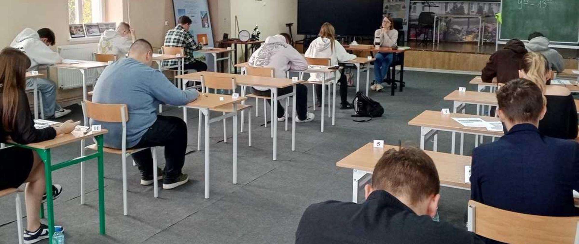 Zdjęcie przedstawia scenę w sali lekcyjnej Zespołu Szkół Centrum Kształcenia Rolniczego im. Kazimierza Wielkiego w Kowalu. W pomieszczeniu panuje atmosfera koncentracji, sugerująca trwający egzamin.
Na pierwszym planie, po prawej stronie, siedzi dziewczyna z długimi blond włosami, ubrana w jasny sweter. Jest pochylona nad ławką, na której leżą dokumenty. Obok jej krzesła stoi czarny plecak marki Puma.
Po lewej stronie, bliżej obiektywu, widać plecy chłopaka w szarej bluzie z kapturem i jasnych spodniach, również skupionego na zadaniu. Przy jego biurku na podłodze leży plastikowa butelka z wodą.
Wokół rozmieszczeni są inni uczniowie, siedzący pojedynczo przy ławkach, wszyscy zajęci pisaniem lub czytaniem.
Tło stanowią ściany w kolorze beżowym. Na środku, nad tablicą multimedialną, widnieje napis "ZESPÓŁ SZKÓŁ CENT[RUM]". W głębi sali, po prawej stronie, znajdują się dwa duże okna wpuszczające światło dzienne. Widoczny jest też stół do piłkarzyków i inne panele wystawowe. Na tablicy kredowej widnieje napis "9-13.", co może wskazywać na godziny trwania zajęć lub egzaminu.