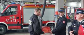 Komendant wręcza strażakowi czerwoną teczkę. Obok niego stoi drugi strażak w mundurze. W tle samochód pożarniczy. 