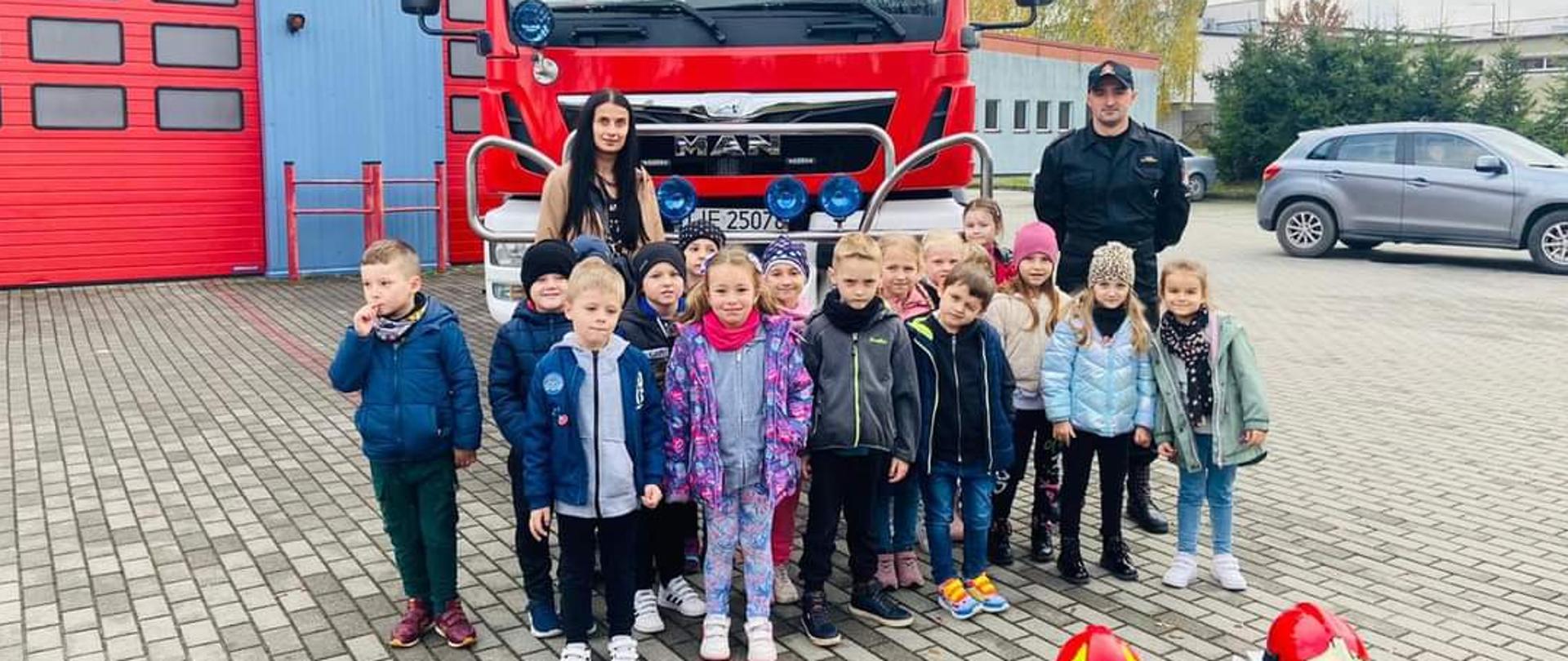 Zdjęcie przedstawia dzieci na tle samochodu ratowniczo-gaśniczego wraz z opiekunem i strażakiem w umundurowaniu dowódczo-sztabowym. Fotografia wykonana przed jednostką ratowniczo-gaśniczą.