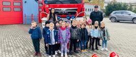 Zdjęcie przedstawia dzieci na tle samochodu ratowniczo-gaśniczego wraz z opiekunem i strażakiem w umundurowaniu dowódczo-sztabowym. Fotografia wykonana przed jednostką ratowniczo-gaśniczą.
