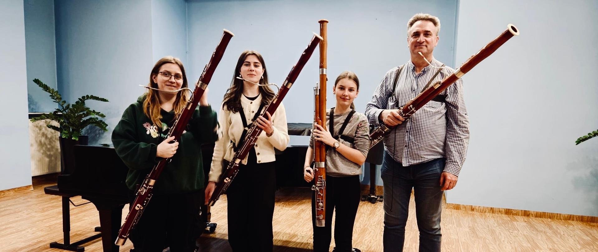 Kolorowe grupowe, zdjęcie. W rzędzie stojące 4 osoby z fagotami na scenie a za nimi czarny fortepian