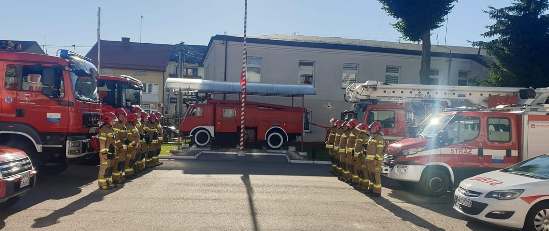 Strażacy stoją naprzeciw siebie podczas zmiany służby. Za nimi stoją samochody pożarnicze. Na środku zabytkowy samochód pożarniczy oraz maszt z flagą z czarnym kirem opuszczoną do połowy.