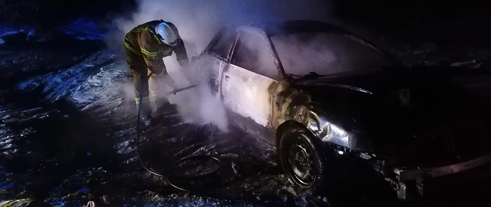 Samochód osobowy po pożarze, obok gaszący za pomocą szybkiego natarcia pożar strażak z OSP Mosty. 