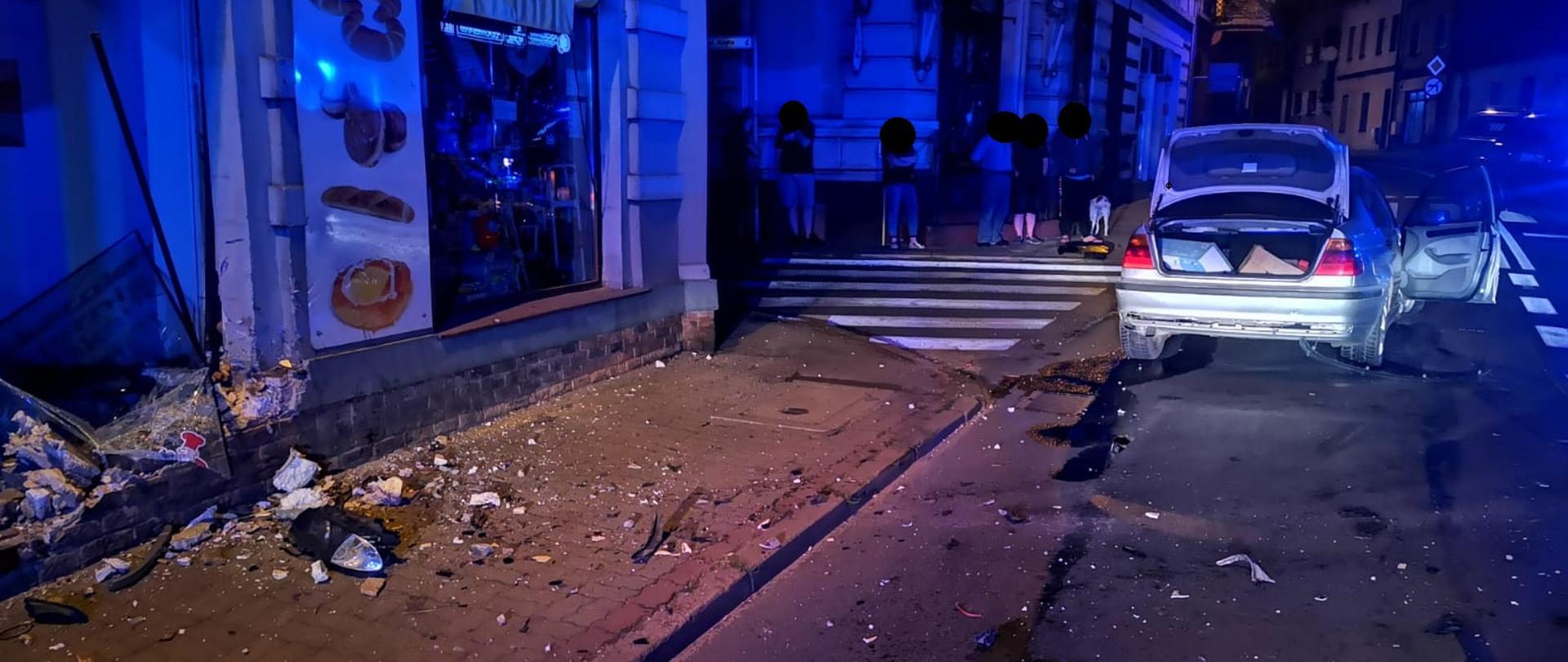 Widać uszkodzoną witrynę sklepu oraz samochód z otwartą klapą bagażnika