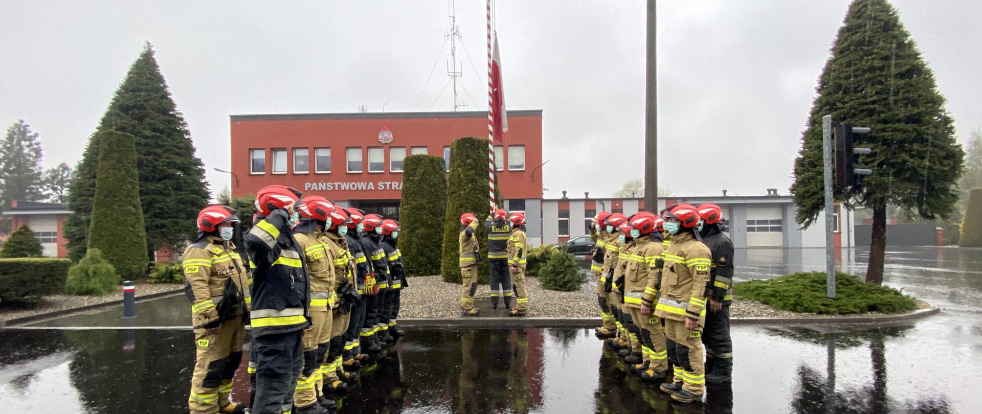 Zdjęcie przedstawia poczet flagowy przed budynkiem komendy PSP, podnoszący flagę na maszt. Obok w szyku zwartym stoją dwie zmiany służbowe w ubraniach specjalnych i maseczkach ochronnych.