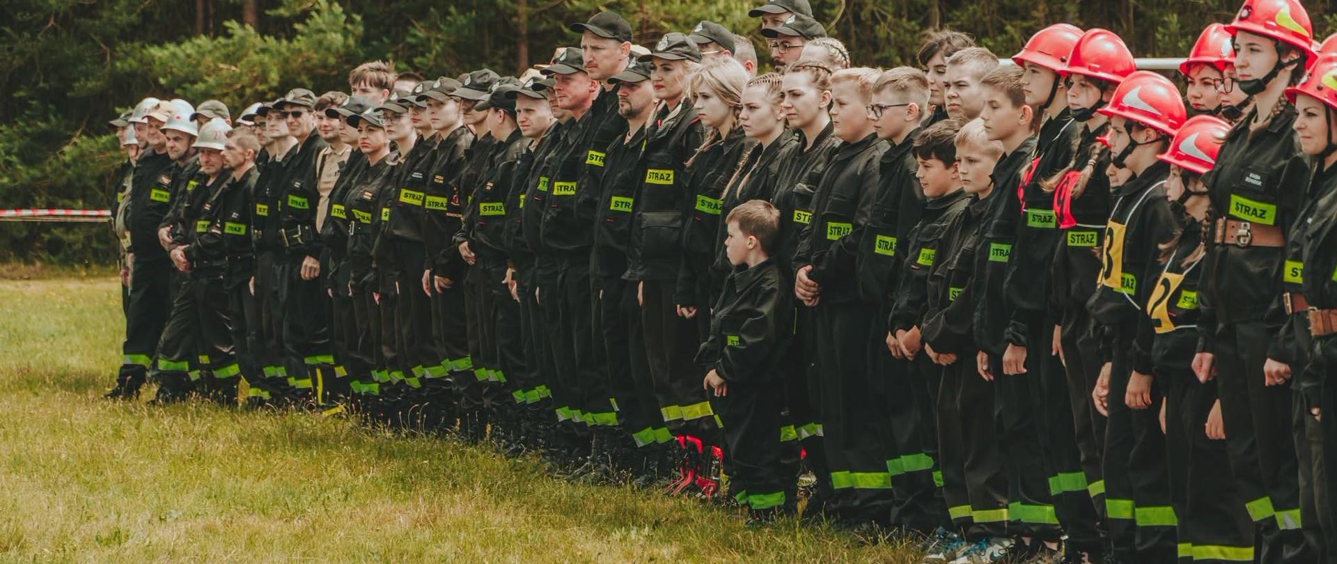 Strażacy z Ochotniczej Straży Pożarnej ustawieni w długim szeregu na trawiastym boisku podczas uroczystej zbiórki przed zawodami. W szeregu stoją zarówno dorośli, jak i młodzież oraz dzieci, wszyscy w czarnych mundurach z odblaskowymi elementami i naszywkami STRAŻ. Część osób ma czarne czapki z daszkiem, a po prawej stronie zawodnicy noszą czerwone hełmy strażackie i numery startowe. W tle widać las w słoneczny dzień.