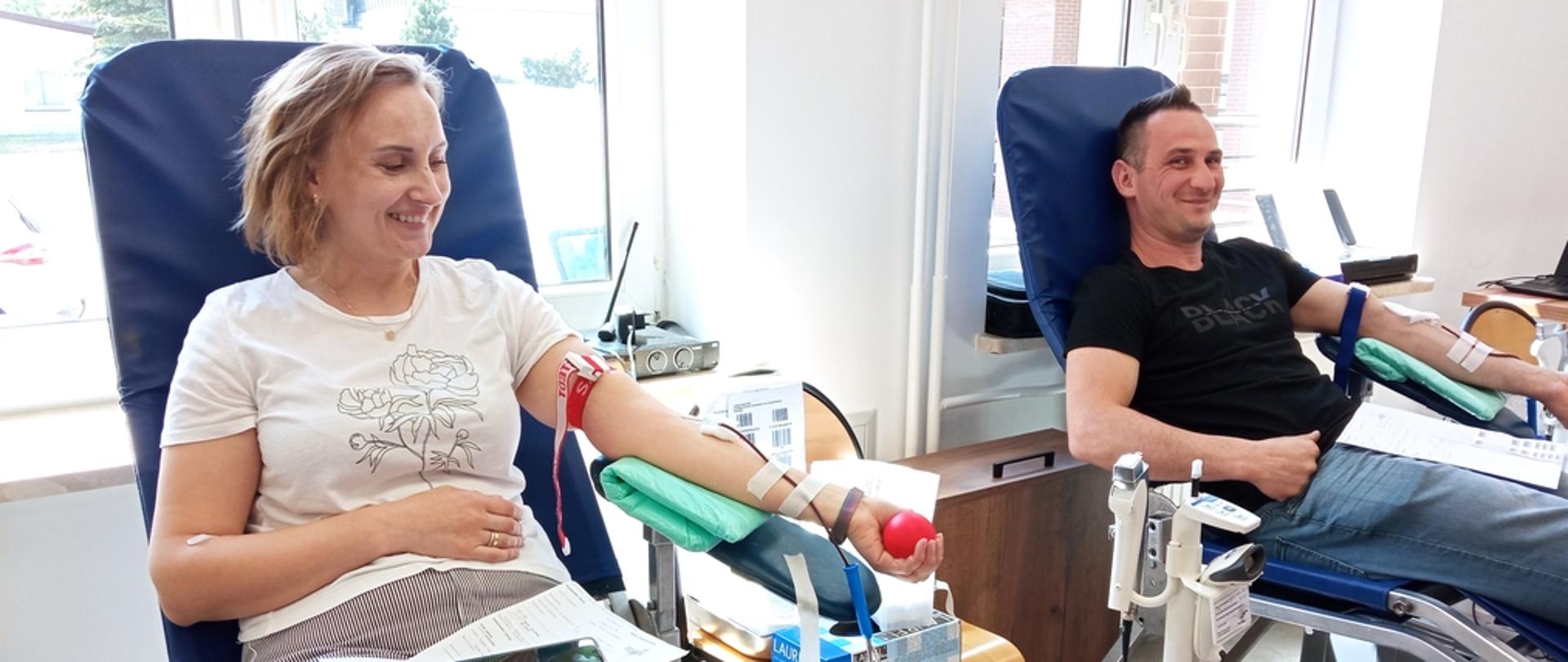 Na zdjęciu widoczni kobieta w białej koszulce i spodniach w paski biało niebieskie i mężczyzna w czarnej koszulce i spodniach dżinsowych oboje siedzą na fotelach koloru niebieskiego i oddają krew w ramach akcji krwiodawstwa w budynku Komendy Powiatowej Państwowej Straży Pożarnej w Gorlicach. 