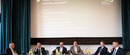 Na scenie sześciu mężczyzn siedzi w fotelach podczas panelu dyskusyjnego II Konferencji Naukowej KRUS - strategia, zdrowie i bezpieczeństwo polskiej wsi. W tle widoczny tytuł konferencji oraz imiona, nazwiska oraz stanowiska panelistów.