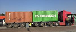Na zdjęciu widać dużą ciężarówkę przewożącą dwa kontenery transportowe.
Po prawej stronie znajduje się czerwony ciągnik siodłowy (kabina ciężarówki).
Za nim stoi zielony kontener z napisem „EVERGREEN”. Dalej z tyłu znajduje się brązowy kontener z napisem „TRITON”. Cały zestaw stoi na utwardzonym parkingu. Pod naczepą pracuje osoba w odblaskowej kamizelce, która coś sprawdza. Obok kabiny ciężarówki stoi druga osoba, zwrócona w stronę pojazdu. W tle widać inne pojazdy ciężarowe oraz drzewa. 