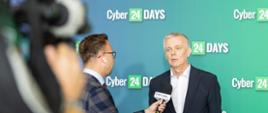 Tomasz Siemoniak, ubrany w granatowy garnitur i białą koszulę, udziela wywiadu dziennikarzowi TVP Info podczas wydarzenia „Cyber 24 Days”. Dziennikarz w okularach i marynarce w kratę trzyma mikrofon skierowany w stronę rozmówcy. Po lewej stronie kadru widoczna jest kamera telewizyjna i rozmyta sylwetka operatora. W tle znajduje się turkusowa ścianka z powtarzającym się logo „Cyber 24 Days”.