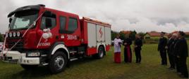 Zdjęcie przedstawia nowy samochód strażacki, który przystrojony jest wstęgą oraz balonami. Ksiądz święci pojazd. Obok stoją druhowie oraz wójt gminy. 