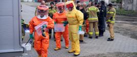 Na pierwszym planie trzech uczestników szkolenia w zakresie przeciwdziałania zagrożeniom CBRNE ubranych w specjalistyczny kombinezon chemiczny, trzymający w dłoniach plastikowe pojemniki, w tle stojący funkcjonariusze PSP