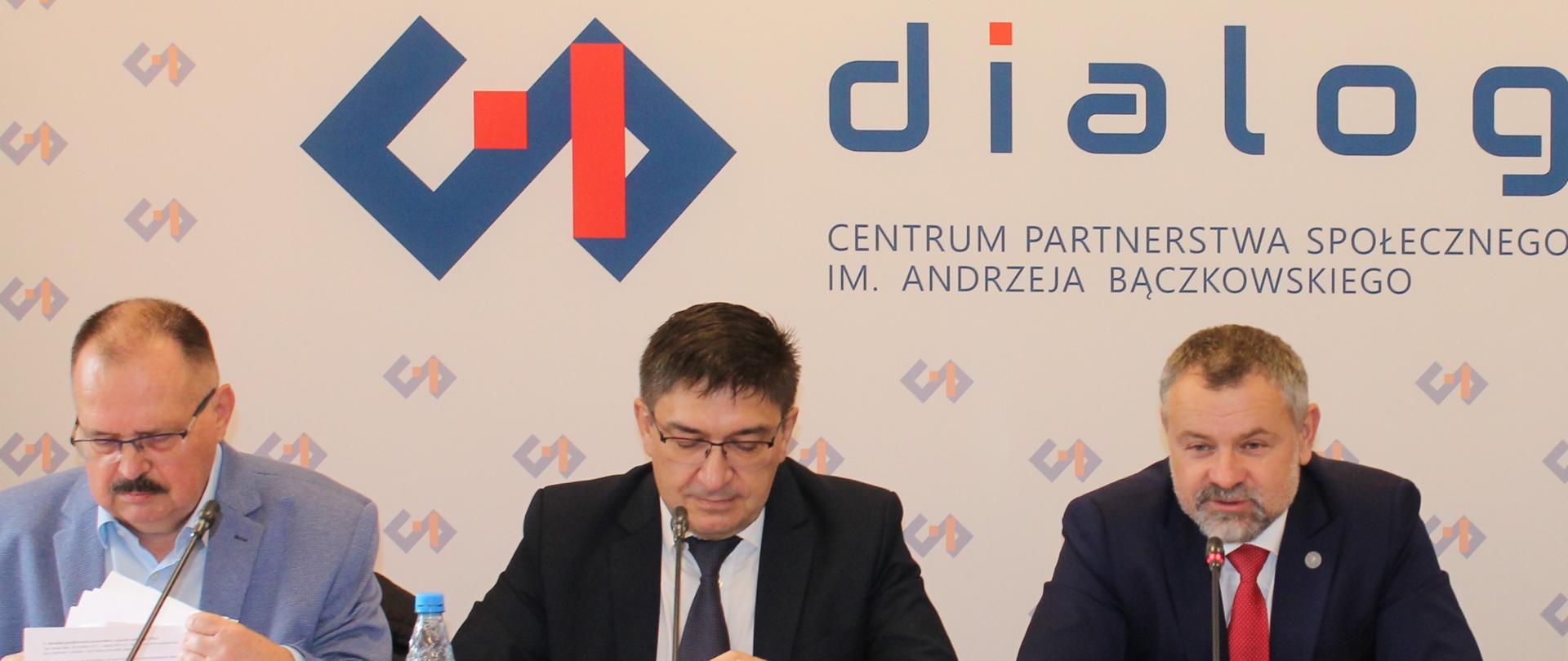 Zdjęcie przedstawia stół prezydialny za którym siedzą: Pan Jerzy Wiertelak, Współprzewodniczący Zespołu Trójstronnego ds. Branży Energetycznej ze strony związków zawodowych; Pan Wojciech Ilnicki, Współprzewodniczący Zespołu Trójstronnego ds. Branży Węgla Brunatnego ze strony związków zawodowych oraz Pan Grzegorz Wrona, Podsekretarz stanu w Ministerstwie Aktywów Państwowych, Współprzewodniczący obu Zespołów ze strony rządowej. W tle znajduje się baner z napisem Centrum Partnerstwa Społecznego „Dialog” im. Andrzeja Bączkowskiego
