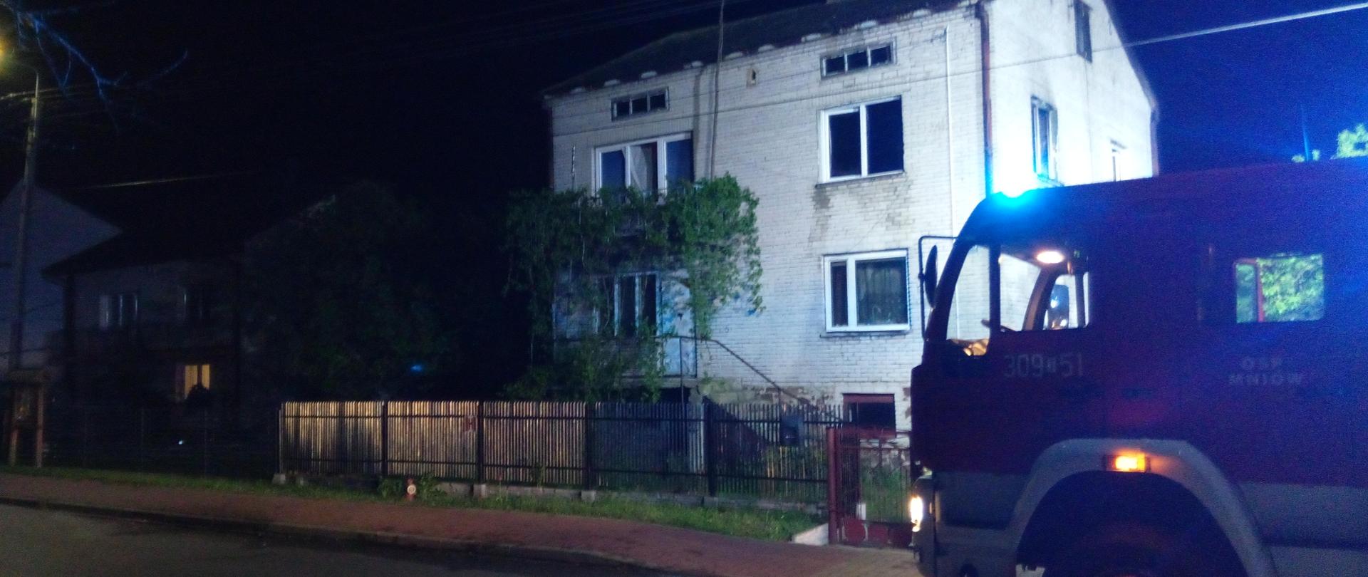 Fotografia przedstawia budynek mieszkalny, w którym wybuchł pożar. Przed budynkiem stoi zaparkowany samochód pożarniczy.
