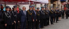 Zdjęcie przedstawia strażaków oraz gości stojących pod trybuną honorową