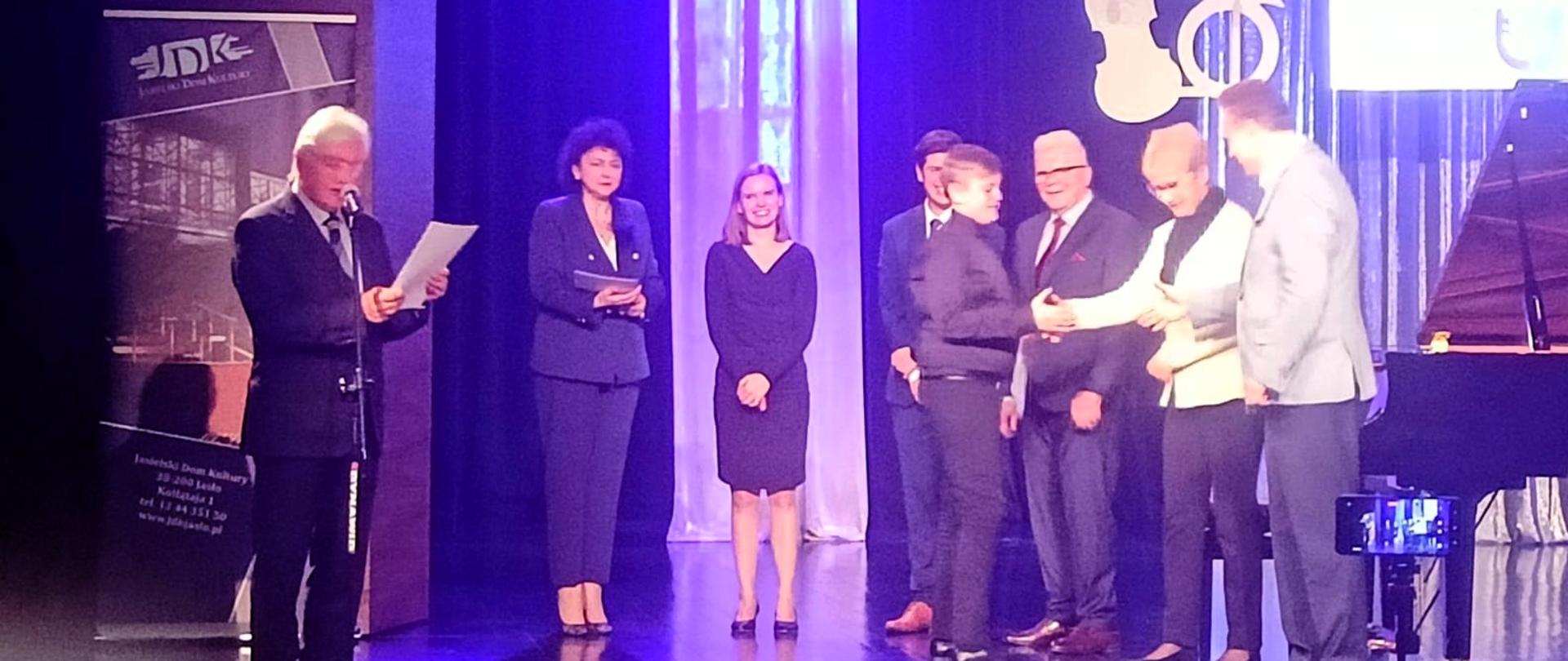 Uroczystość wręczenia nagród na scenie. Od lewej strony, mężczyzna z siwymi włosami i w ciemnym garniturze przemawia do mikrofonu, trzymając kartki. Za nim widoczne są zasłony i logo na sztaludze z tekstem. Pośrodku sceny stoją cztery osoby. Jedna osoba wręcza drugiej nagrodę. Po prawej stronie widać fortepian i stojak z nutami. Po prawej stronie sceny widoczne są instrumenty muzyczne.