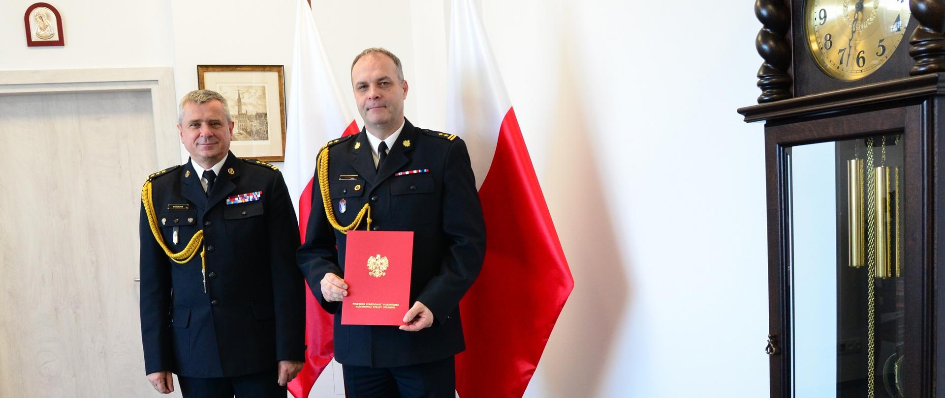 Pomorski komendant wojewódzki Państwowej Straży Pożarnej oraz strażak trzymający akt powołania stoją wspólnie na tle flagi Polski.