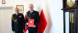Pomorski komendant wojewódzki Państwowej Straży Pożarnej oraz strażak trzymający akt powołania stoją wspólnie na tle flagi Polski.