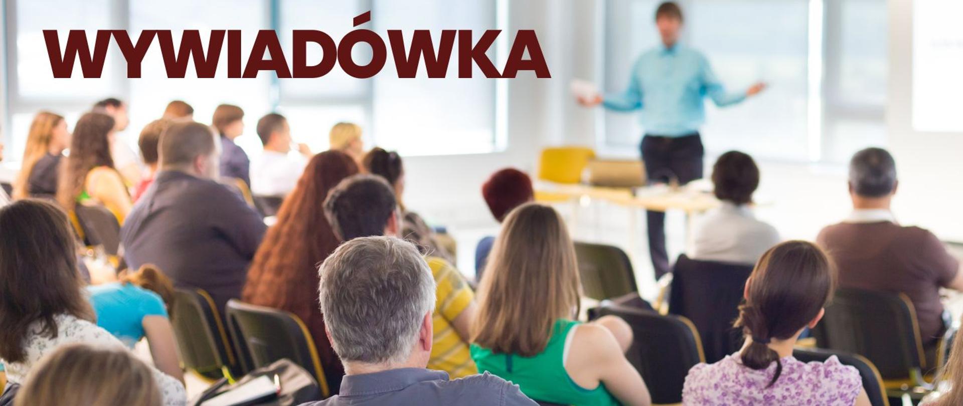 klasa lekcyjna z kilkoma osobami siedzącymi na krzesłach oraz nauczyciel na tle okien i tablicy z napisem wywiadówka w kolorze bordowym