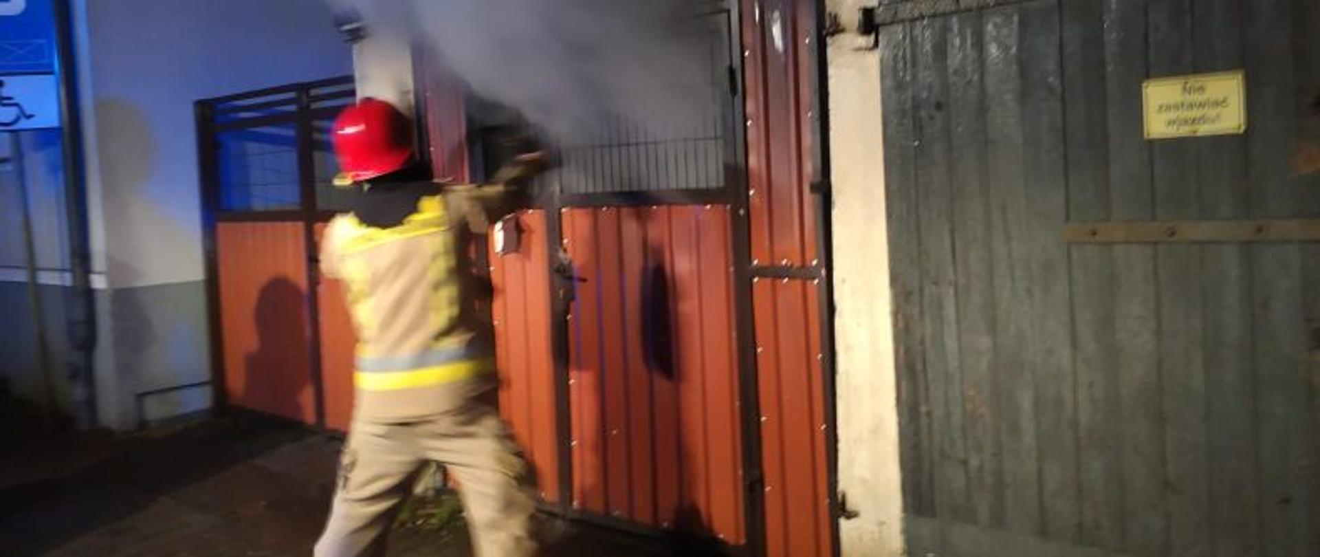 Zdjęcie przedstawia strażaka w piaskowym mundurze i czerwonym hełmie. Strażak w rękach trzyma prądownicę i kieruje strumień wody do środka wiaty. Ze środka unosi się dużo gęstego dymu