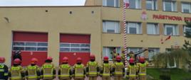 Zdjęcie przedstawia uroczystą zmianę służby z podniesieniem flagi na maszt przed budynkiem KP PSP w Kętrzynie. Strażacy ubrani w piaskowe ubranie specjalne z czerwonym hełmem stoją w linii przed masztem. Trzech strażaków tak samo ubranych stoi w linii przy maszcie. Dwóch strażaków w czarnym mundurze wyjściowym ustawionych prostopadle do strażaków.