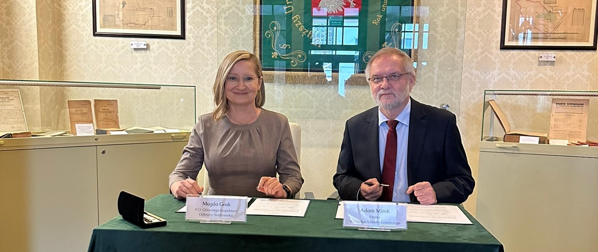 Kobieta i mężczyzna siedzą przy stoliku ubranym w zielony obrus. Mają w rękach długopisy i pozują do zdjęcia.