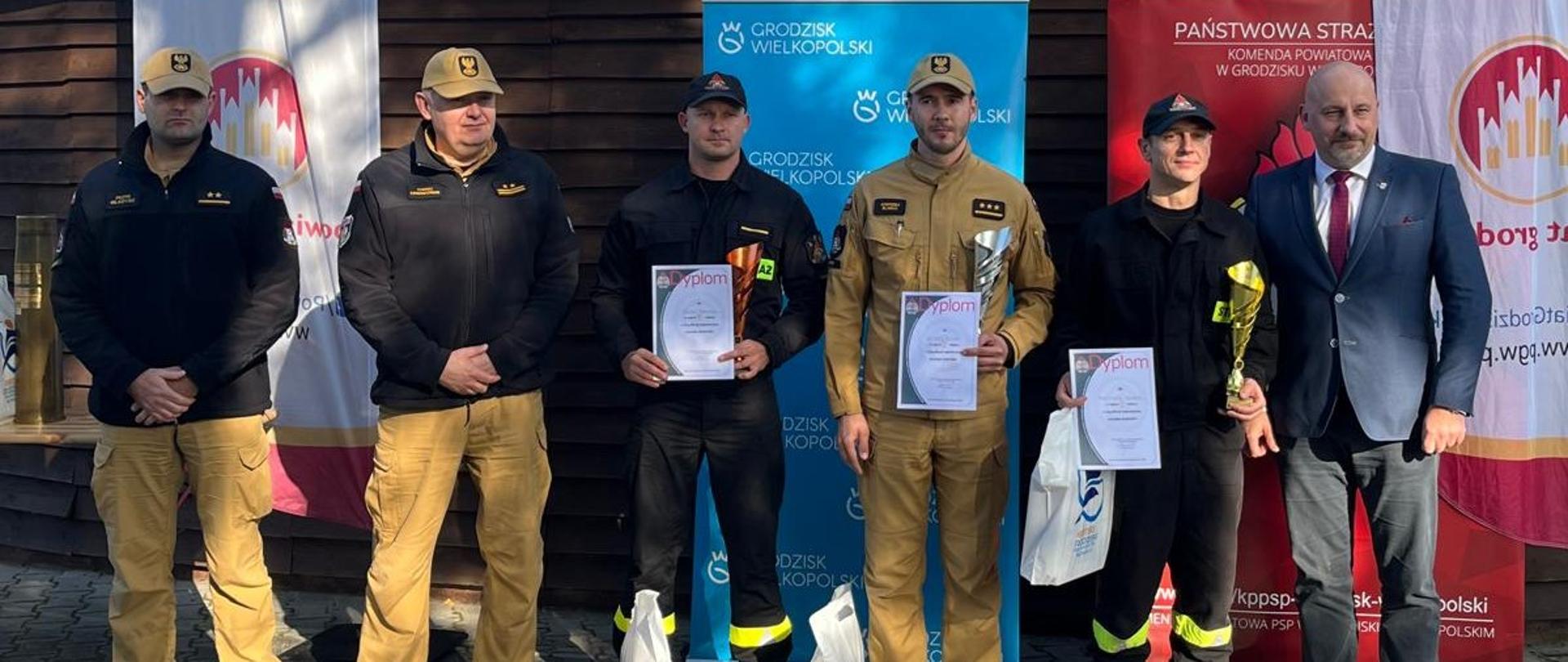 Sześciu mężczyzn stoi w szeregu, część z nich trzyma w ręku dyplomy, pięciu ubranych jest w mundury Państwowej Straży Pożarnej, pierwszy z prawej ubrany jest w garnitur